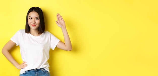 Free photo friendly asian woman in white tshirt waving hand and saying hello greeting you standing over yellow