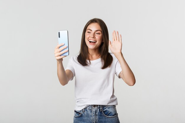 Friendly attractive girl saying hi, waving hand at smartphone during video call, having conversation.