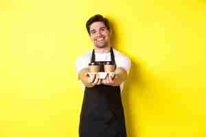 Free photo friendly barista in black apron giving takeaway order, holding two cups of coffee and smiling, standing over yellow background