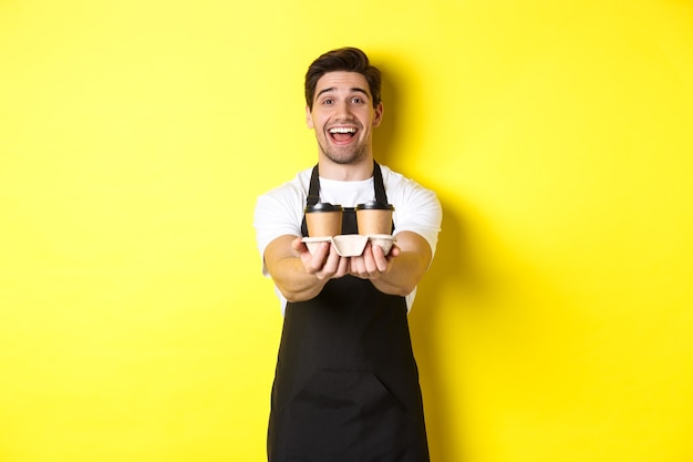 Friendly barista in black apron giving takeaway order, holding two cups of coffee and smiling, standing over yellow background.