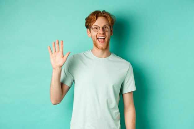 Friendly bearded guy in glasses saying hello, waving hand to greet and welcome you, standing cheerful and smiling over turquoise background.