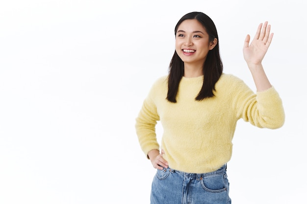 Friendly beautiful asian woman saying hello, smiling happy and waving hand to the left, seeing friend, team leader make goodbye gesture after productive work, feeling pleased and delighted