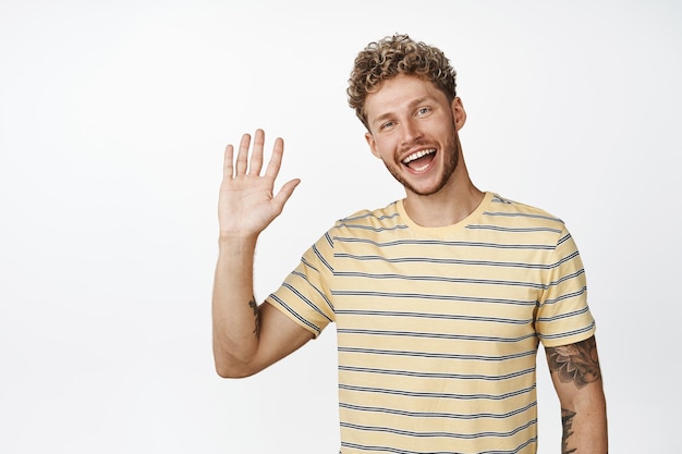 Free photo friendly blond guy saying hello waving hand and smiling at camera standing over white background