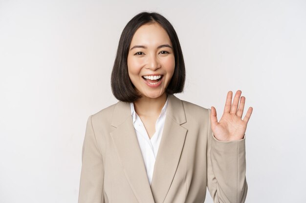 Friendly business woman asian office lady waving hand and saying hello hi gesture standing over white background