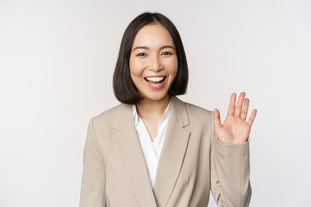 Friendly business woman asian office lady waving hand and saying hello hi gesture standing over white background