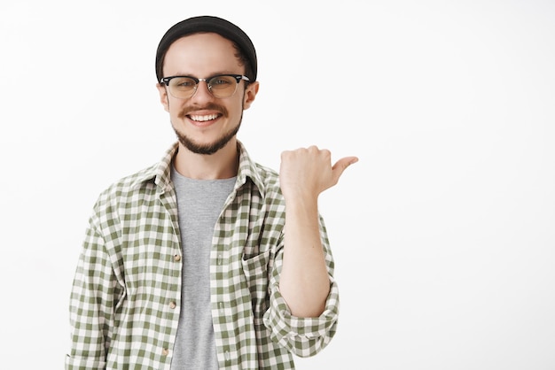 Friendly carefree and satisfied good-looking male customer with beard in glasses black beanie and casual green shirt pointing right with thumb and smiling joyfully giving advice or showing way