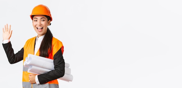 Friendly cheerful asian female construction manager lead architect in safety helmet and jacket carry blueprints documents of building project waving hand to say hello greeting someone