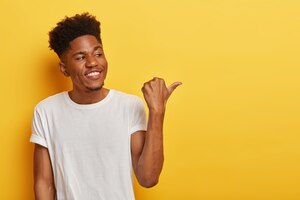 Friendly cheerful dark skinned hipster man discusses interesting copy space and points thumb away on right side, shows some place or direction, has curly trendy haircut, wears casual white t shirt