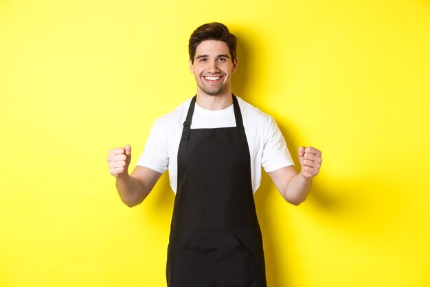 Friendly coffee shop waiter standing with raised hands, place for your sign or logo, standing over yellow background.