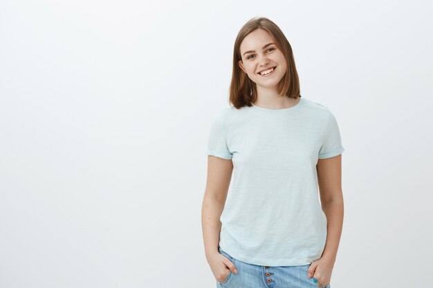 Friendly cute white girl with short haircut and lovely flapped ear smiling joyfully holding hands in pockets and gazing with joyful entertained look posing over white wall