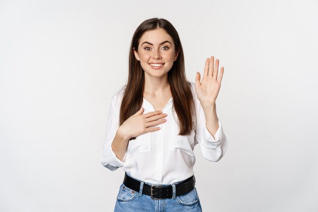 Friendly cute woman raising hand and introduce herself, saying name, pointing at her, saying hello, standing over white background