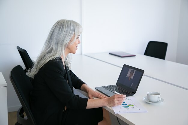 커피 한잔과 함께 테이블에 앉아 다이어그램을 분석하는 동안 노트북에서 화상 채팅을 통해 동료에게 말하는 친절한 여성 회사원. 온라인 커뮤니케이션 개념