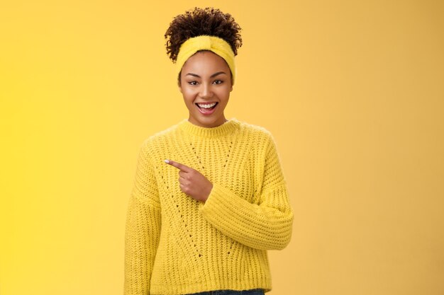 머리띠 스웨터를 입은 친절하고 잘 생긴 열정적인 흑인 매력적인 소녀가 활짝 웃으며 노란색 배경을 가진 완벽한 빈 공간 광고를 보여주는 멋진 장소를 가리키고 있습니다.