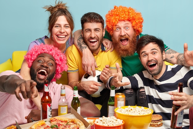 Friendly happy four young men and one woman embrace and look joyfully at screen of TV, enjoy watching television and funny film, hold ball for football, have fun at home.