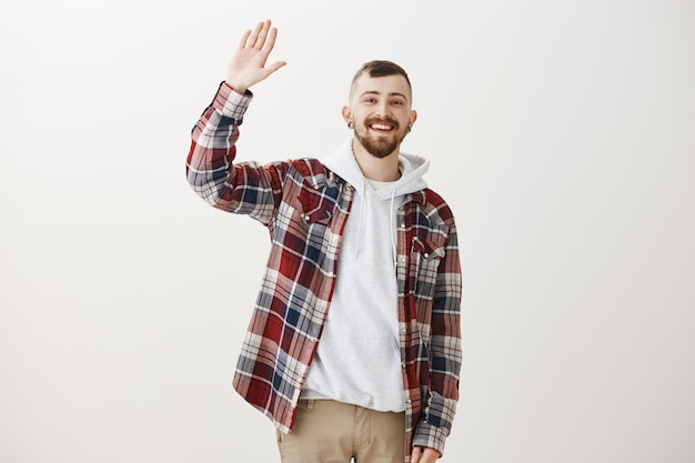 Friendly happy hipster guy waving raised hand to say hello, greeting you