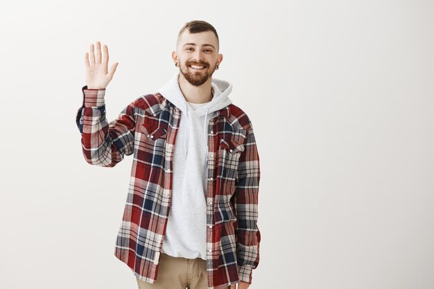 Friendly happy hipster guy waving raised hand to say hello, greeting you