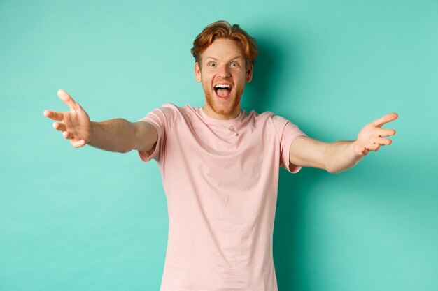 Friendly and happy young man with ginger hair, stretch out hands in warm welcome, reaching for hug and smiling joyfully, standing in t-shirt over mint background.