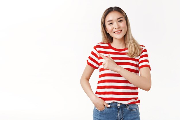 Friendly kind charismatic asian blond girl tilt head smiling pleasantly carefree speaking discuss interesting copy space ad pointing left index finger showing friend place direction white background