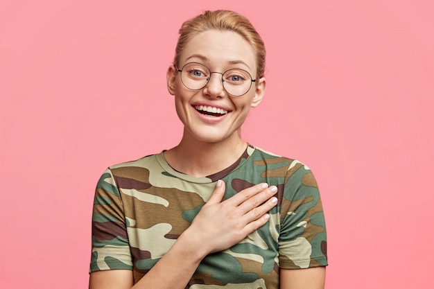 Donna bionda attraente dall'aspetto amichevole che viene toccata con una storia che penetra nel cuore, ha un sorriso piacevole, tiene la mano sul petto, isolata sul rosa