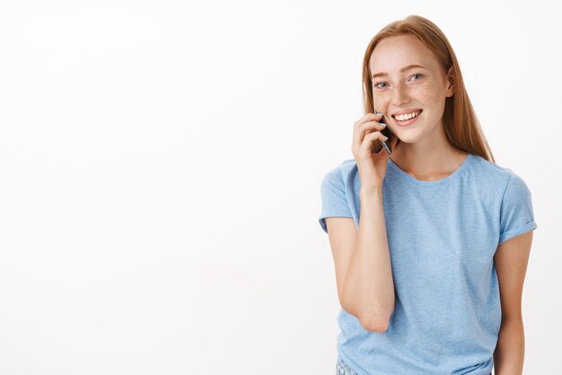 빨간 머리와 주근깨가있는 친절하고 매력적인 사교적 인 여성이 회색 벽 위에 즐겁고 편안하게 전화를하는 동안 귀 근처에 핸드폰을 들고 스마트 폰으로 이야기합니다.