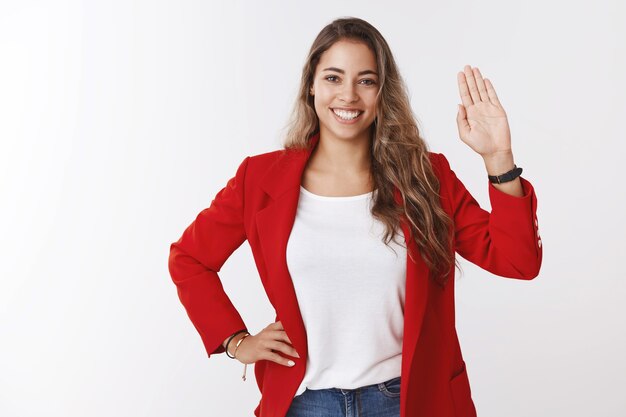 Friendly-looking confident attractive young european curly-haired 25s woman wearing red jacket waving raised palm hello welcome gesture smiling, greeting team members, saying hi meeting new people