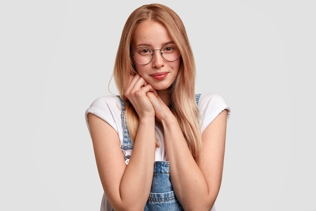 Friendly looking European woman youngster with appealing look, keeps hands together near chin, wears spectacles and denim dungarees