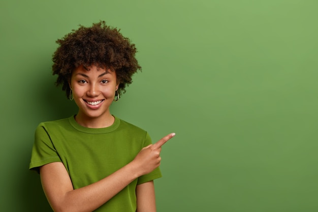 Free photo friendly looking glad female shop assistant happy to help customers, shows way and demonstrates discounts in shop, points fore finger aside on empty space over green wall.