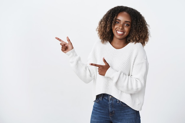 Дружелюбная живая приятная афро-американская женщина-коллега в белом свитере с довольной улыбкой и указательным пальцем влево