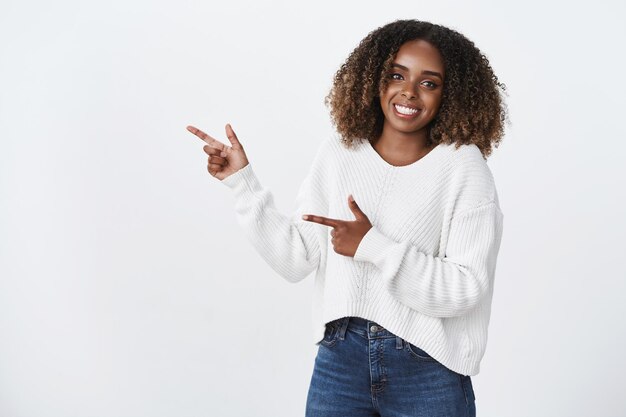 Дружелюбная живая приятная афро-американская женщина-коллега в белом свитере с довольной улыбкой и указательным пальцем влево