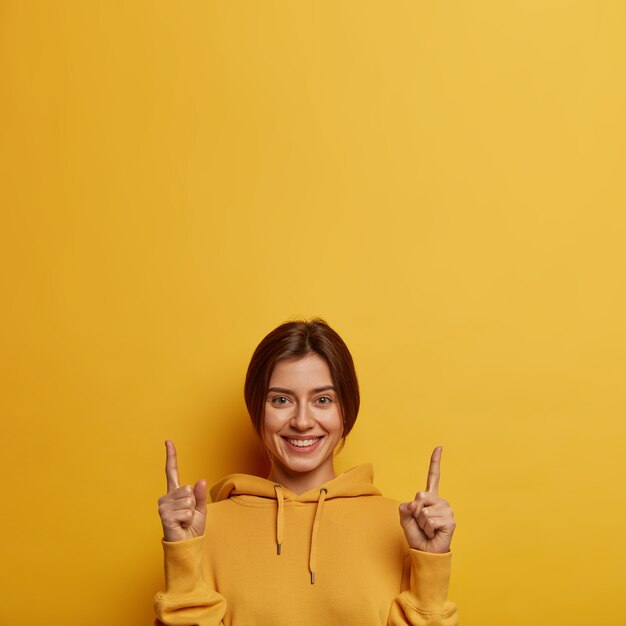 Friendly looking lovely woman shows promo with joy, points above with both index fingers, gives recommendation or advice, wears yellow hoodie