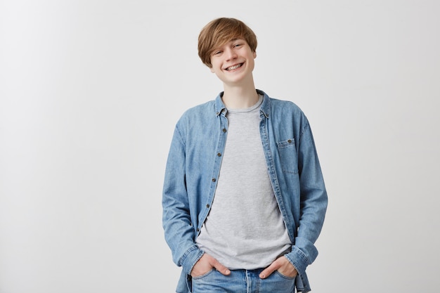 Friendly looking positive european young man with fair hair and blue eyes, smiling broadly with braces, during nice conversation with friends, laughing at jokes, standing .