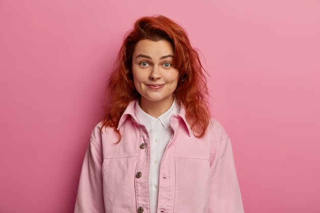 Friendly looking tender girl has red wavy hair, smiles with dimples on cheeks, looks carefree and relaxed, being self assured, isolated over pink space. People