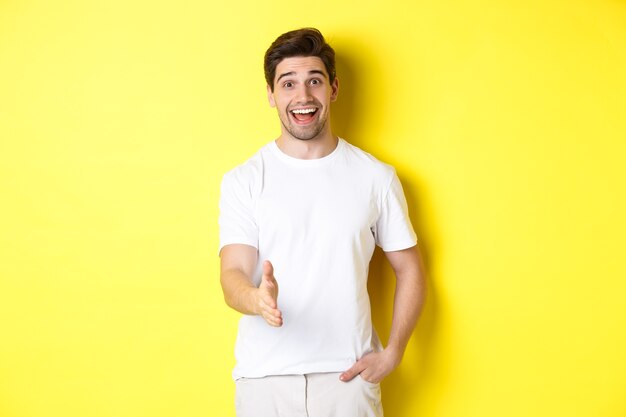 Friendly man greeting you with handshake, smiling amused, saying hello, standing over yellow