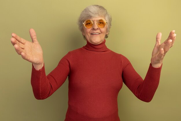 Friendly old woman wearing red turtleneck sweater and sunglasses meeting guests with wide open arms doing i am glad to see you gesture isolated on olive green wall