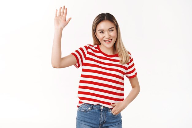 Friendly outgoing sociable cute asian blond girl raise hand waving palm hi hello gesture smiling broadly happy introduce herself new members, standing white wall