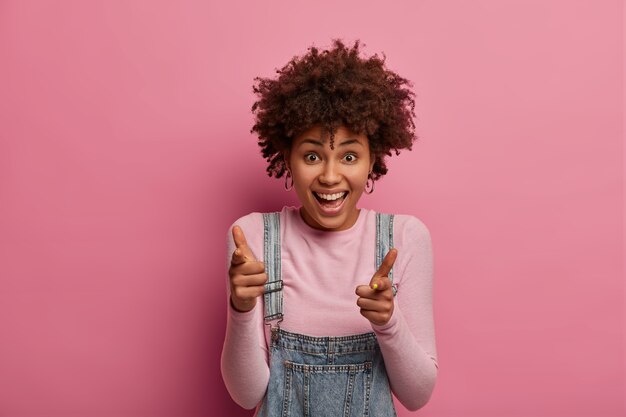 Friendly outgoing woman makes finger pistols gesture as praising you for good job, congratulates groupmate with successfully passed exam and achievement, laughs sincerely, poses in pink .