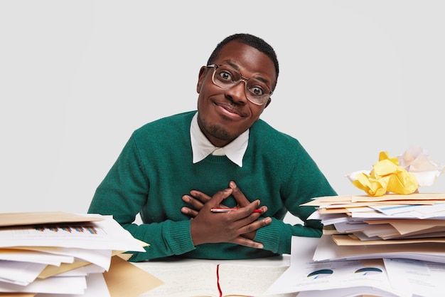 Friendly positive black man keeps hands on chest, holds pen, has pleased facial expression, being thankful to friend for helping with paper work