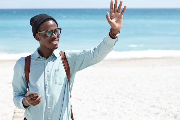 Friendly positive smiling young Afro American man in trendy hat and shades holding smartphone and waving hand, hailing friends while having walk on city beach