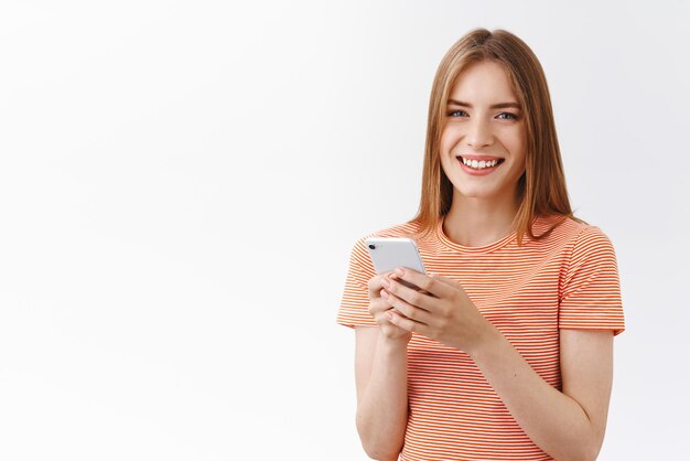 Дружелюбная симпатичная молодая беззаботная женщина в полосатой футболке держит смартфон, улыбается и смотрит в камеру, в восторге от просмотра интернета, звонит по телефону, пишет забавную историю, стоит на белом фоне