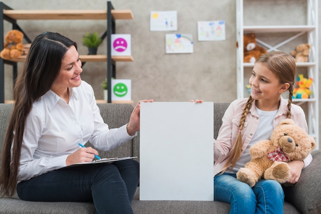 Free photo friendly psychologist and little girl holding blank placard sitting on sofa