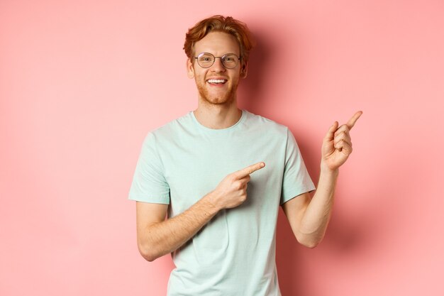 Friendly redhead guy in glasses pointing at upper right corner showing promo banner and smiling at c...