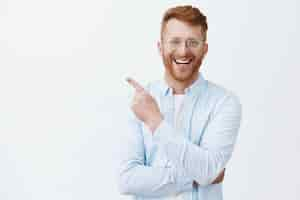 Free photo friendly redhead man with bristle in transparent glasses and striped shirt pointing at upper left corner, smiling broadly with satisfied and pleasant look