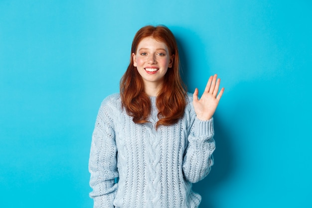Friendly redhead teenage girl saying hi, waving hand in hello gesture and smiling, standing against blue background