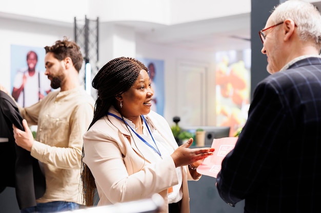 Foto gratuita cordiale e sorridente assistente alle vendite di una donna afroamericana che spiega le regole dello shopping del black friday nel negozio di abbigliamento al cliente senior. operaio al dettaglio che tiene il volantino di vendita che parla con l'acquirente