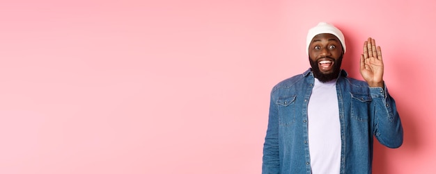 Free photo friendly smiling black man saying hello waving hand greeting you standing over pink background