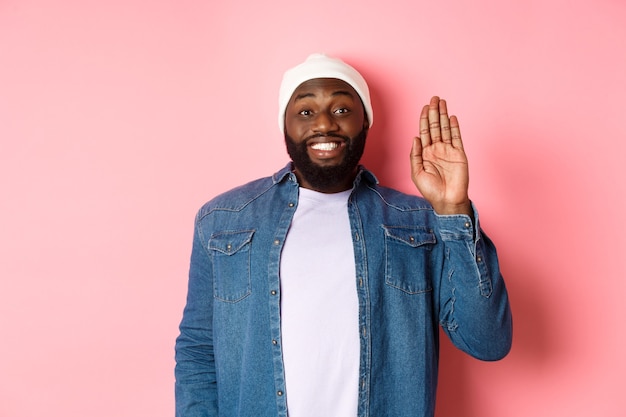 Friendly smiling Black man saying hello, waving hand, greeting you, standing over pink background