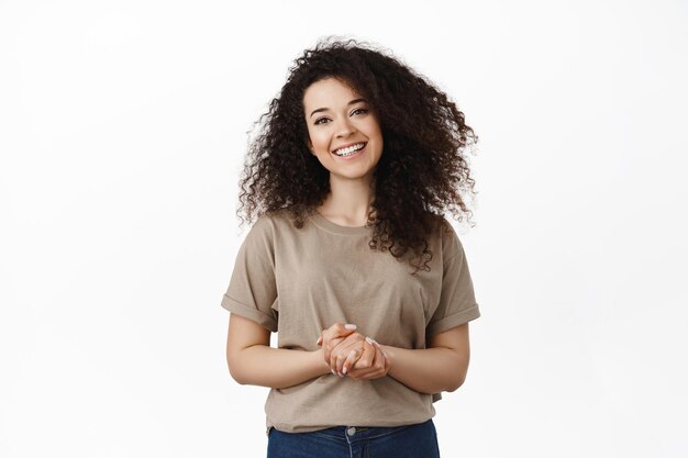 Friendly smiling brunette woman ready to help and assist, holding hands together and looking pleasant, standing in t-shirt against white background.