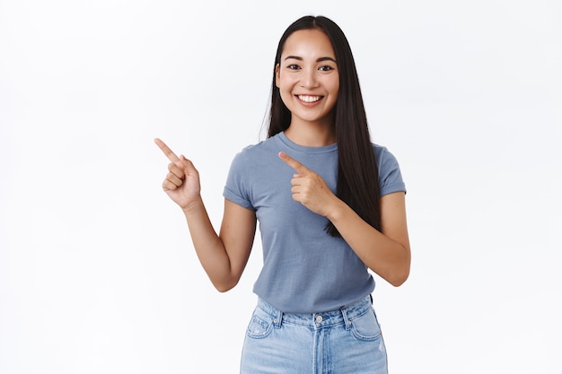 Friendly, smiling happy asian girl give advice where find what need, pointing fingers left, grinning carefree, recommend advertisement, promote product, discuss new store, white wall