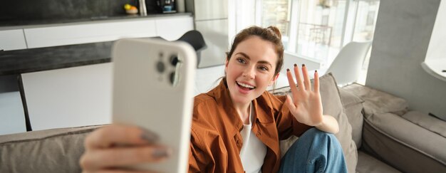 friendly smiling young woman sitting on couch and waving hand at smartphone camera video chats