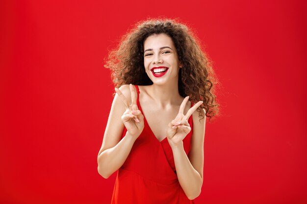 Friendly sociable goodlooking happy girl with curly hairstyle wearing elegant red dress and evening ...